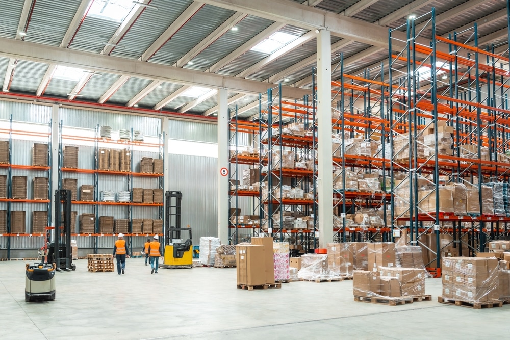 Binnenzicht van groot magazijn met pallets, heftrucks en medewerkers in oranje hesjes